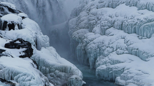 A photo of the base of a waterfall. Falling water can be seen in the top left of the image, and there is an open channel with flowing water between two impressive ice walls. On the left, a few dark rocks are visible between the frozen areas but otherwise every surface is covered. The water has solidified into long fingers of cyan icicles and the tops have a deep coating of snow.