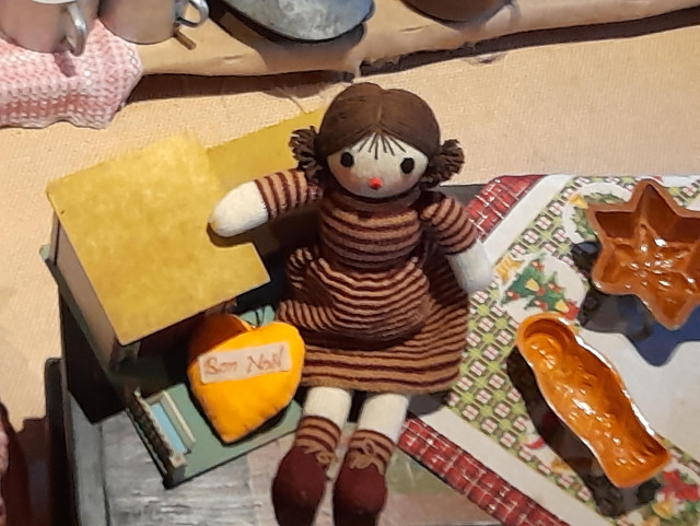 A knitted doll in a striped knitted dress leans against a doll's house. She has brown rat tails, a red scrunchy nose and round dark eyes. Next to it are baking moulds made of clay.