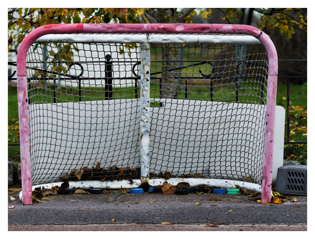 En ishockeybur med olikfärgade plastpuckar. Buren är solblekt röd med vita innerstolpar och svart nät. I bakgrunden hänger höstlöv över ett svart järnstaket och en del av ett vit hus syns.