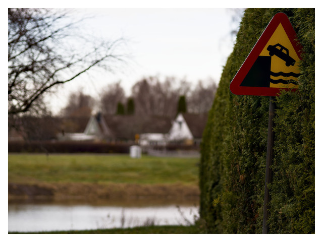 Uppe till höger syns en vägskylt, Varning för att köra över kanten ner i vattnet. Skylten sticker delvis in i en grön häck. I den suddiga bakgrunden syns ett litet vattendrag och på andra sidan några villor.