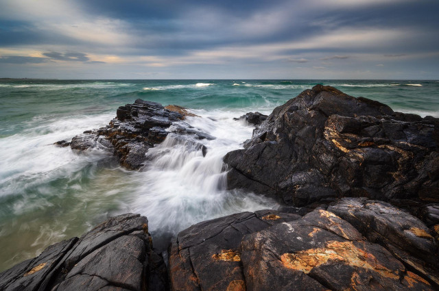 an evening seascape with dramatic skies and black and orange rocks with some water flowing over the rocks from the incoming wave