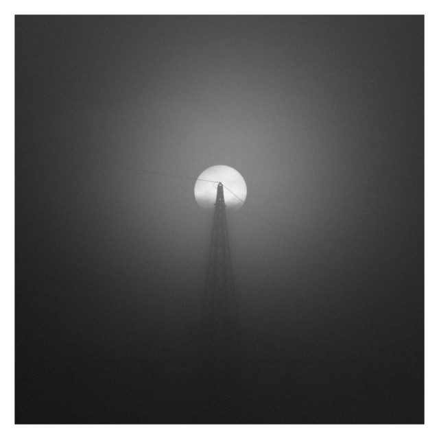 This is a square black-and-white image with a white frame, capturing a foggy scene with a power line tower silhouetted in the foreground. The tower, centered in the image, rises up to meet the bright, round disk of the sun, which appears to be positioned directly behind the top of the tower. The sun, diffused through the fog, creates a soft glow, emphasizing the hazy atmosphere. Power lines stretch outward from the top of the tower, faintly visible against the fog. The overall scene has a surreal, almost otherworldly quality due to the fog, minimal details, and the stark contrast between the dark tower and the bright, softened sun. The background fades into varying shades of gray, leaving no distinguishable horizon or landscape, which enhances the sense of isolation and calm.