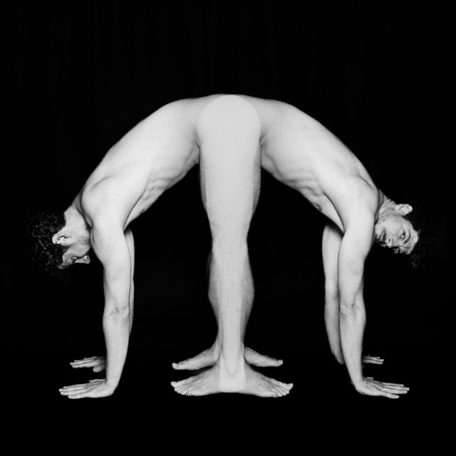 A naked man is standing with his sides to the camera, legs straight and arms flat on the ground on a dark background. There are two superimposed images of him facing right and left. In one of the images, his face looks at the camera. Black and white. 