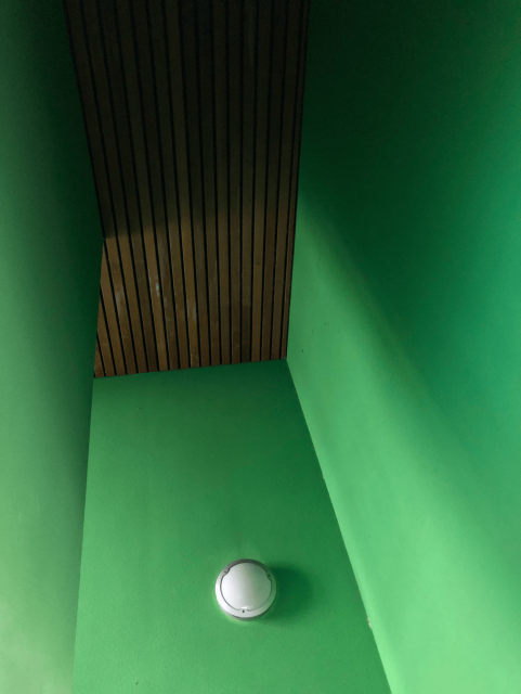 Three green walls and a brown stained wooden ceiling. On the center wall, near the bottom of the frame, is a round white wall lamp.