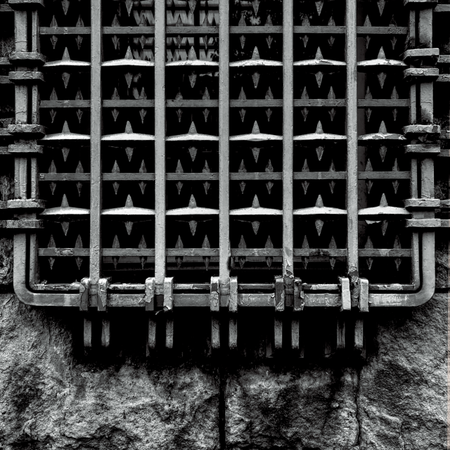 A black and white photo of old iron bars with stylized spikes covering a window embedded in a coarse stone wall.