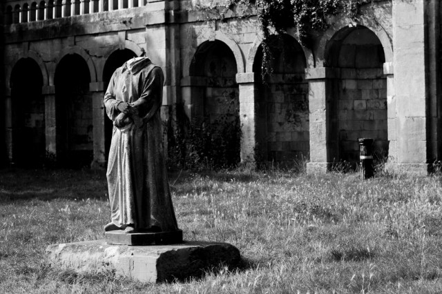 Monochrome photo of a statue taken from the front and to the left. It stands on a rough stone plinth in grassy area a short distance from a brick wall with arched alcoves. The statue is of a standing figure wearing a long gown with their right hand resting on their left wrist resting on their waist. The statue has no head.