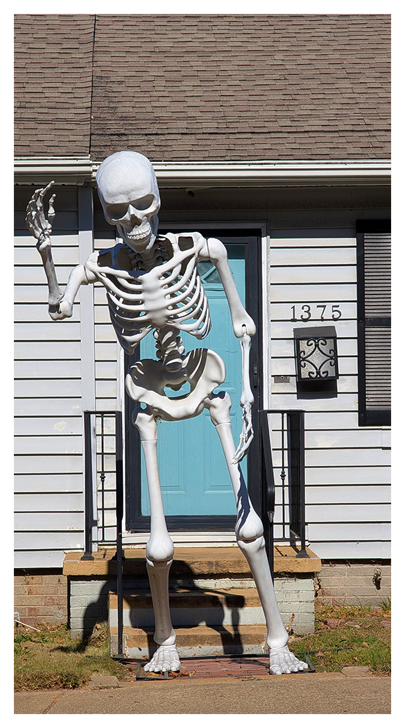 daytime. a larger-than-human sized skeleton stands in front of a single-story house appearing to wave.