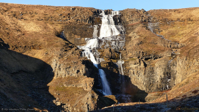 A photo of a white waterfall, dropping down a honey-coloured rockface. Strong sunlight from the left is casting shadows across the multiple steps along the way down. The sky above is clear cyan.