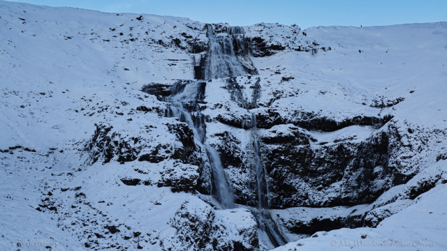 A photo of a waterfall, dropping down an ice-covered rockface. The scene doesn't display any light source although the sky above seems to be clear cyan. Deep snow is piled up on either side of the cascade.