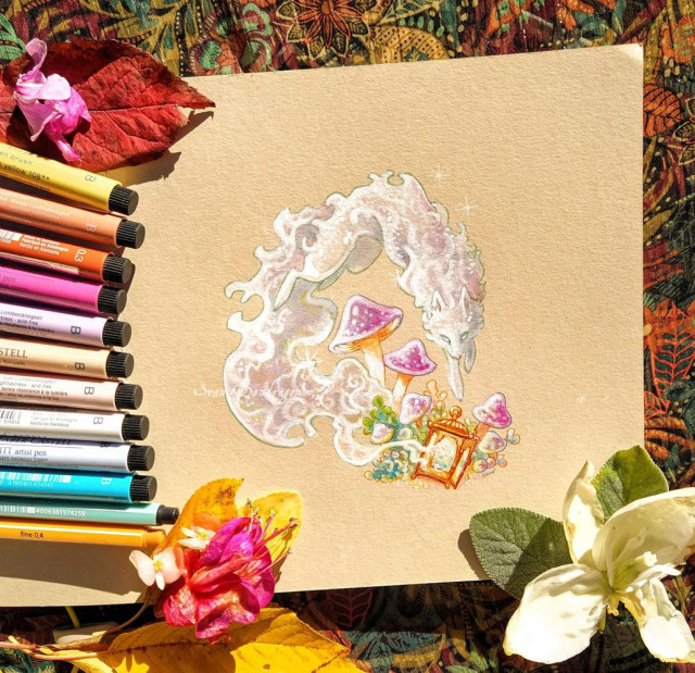 Photo shows a tan paper surrounded by colorfull markers, real leaves and flowers of autumn. On the center of the paper are a drawing of an etheral white shiny fox, jumping outside a lantern, its fur waving like smoke or water. beside the lantern and below the fox are some violet mushrooms. 