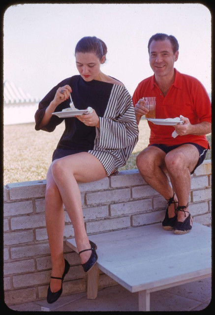 A color photograph featuring a woman and a man seated on what appears to be an outdoor ledge or platform. The woman, dressed in a black top with vertical stripes, is holding a plate of food while the man beside her sports a red polo shirt and dark shorts. They both have plates containing food items, possibly from a dining scenario such as a barbecue or picnic setup. In terms of context provided by the reference information, this image captures an atmosphere reminiscent of mid-20th century leisure activities in Florida during December 1954 at La Coquille Club, indicating that it is likely taken outdoors given its bright lighting and casual attire suited for warm weather.