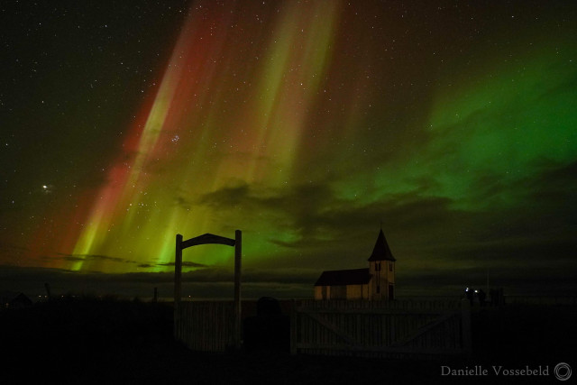 Where do I start. A gate to the church, church in the background, both almost black. The lights from the sky shooting downwards are red and yellow, the wave from left to right is green, . There are some clouds, and a lot of white dots (stars). 