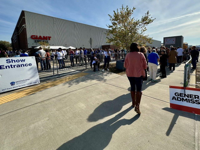 Color photo of people in a long line snaking through a maze of crowd barriers to enter the Giant Expo Hall, Farm Show Center, Harrisburg PA. A brillant blue late October sky, temperature 71F.