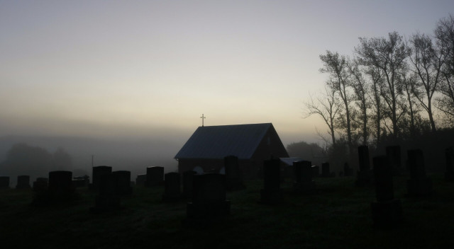 Photograph of a cemetery in the morning fog, with a very pale blue sky, yellow and green hues, as the sun is rising but hidden in the thick fog. A mausoleum building is in the centre, with a metal roof and a cross on top. A row of tall trees is on the right. Graves with their tombstones are all around the building. The picture is taken from behind, as if the building and stones are facing the sun, the village being completely lost in the fog.