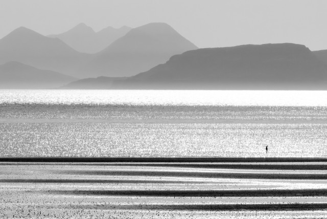 A black and white image of a person in the distance walking alongside the shoreline, with mountains in the distance