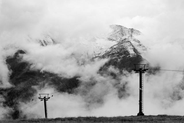 This is a dramatic black and white image of a mountainous landscape partially obscured by fog and clouds. The main focal point is a towering snow-capped peak, its upper reaches visible through the mist. In the foreground, two ski lift towers rise up, their silhouettes contrasting against the hazy, atmospheric scene. The overall image conveys a sense of scale, power, and the mystery of mountain environments shrouded in shifting weather conditions.