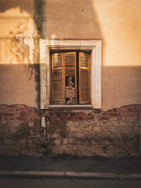 Se puede ver una fotografía de una ventana vieja en la parte central. La mitad inferior y un poco de la superior está en sombra. En esta mitad inferior se ve un muro roido donde asoman ladrillos. En la superior hay un contraste de luz y sombras del atardecer.