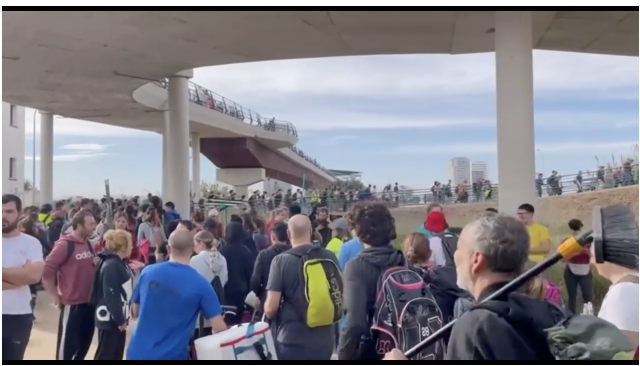 A long line of people threads its way over and under large overpasses. Many are holding brooms, wearing backpacks, or carrying other tools. 