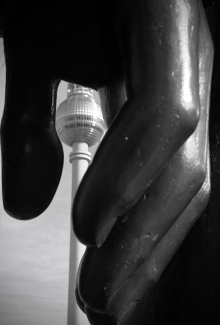 A close-up of a large black sculpture of a hand, with the Berlin TV Tower partially visible in the background. The image is in black and white.