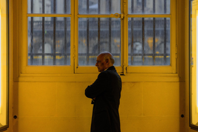 A silhouetted profile view of an elderly man in a dark coat standing against a bright yellow wall. Large windows with yellow frames fill the background, creating a stark contrast between the man's dark figure and the vibrant surroundings. The composition suggests a mood of contemplation or solitude.