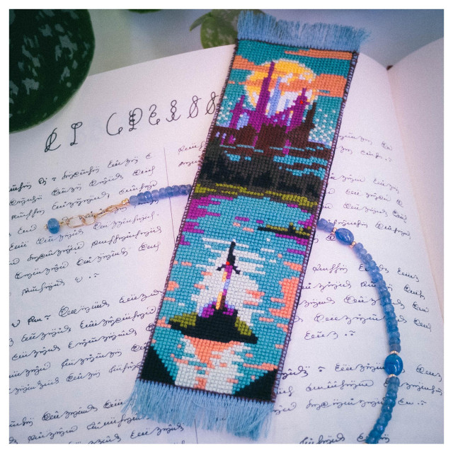 A cross stitched bookmark on a book written in gibberish. The bookmark depicts a fantasy castle in pink tones, against a full moon at the very top. At the bottom, there is Excalibur on a rock. The colors are very saturated, dominated by blue-greens, magenta and orange

