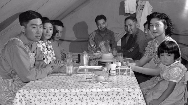 A Japanese family sitting at a table in a tent during the internment of Japanese Americans. 8 people in total in photo, one woman in the back my cousin’s mother doesn’t even look at the camera. She looks distraught.  My aunt, a child then sits in the front. Tin table wear, glass water cups and a box of Mickey Mouse Cookies sits on the table in front. 