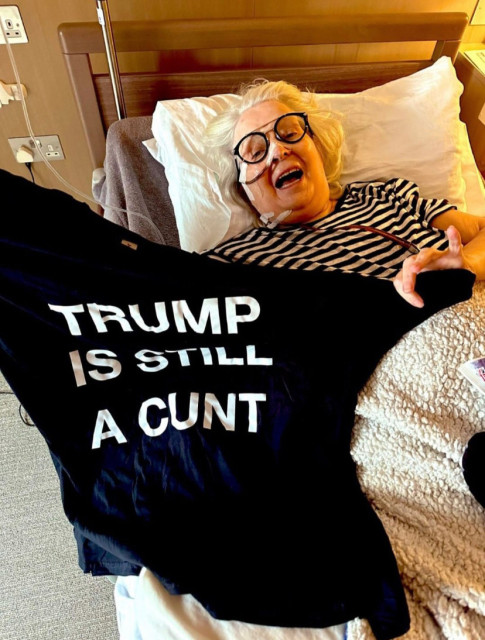 Comedian Janey Godley is lying in bed, intubated. She is wearing a black and white striped shirt and black round-frame glasses, and is under a wool blanket. She has an open-mouthed smile and is holding up a black T-shirt with bold, white sans-serif lettering which reads: "Trump is Still a Cunt".