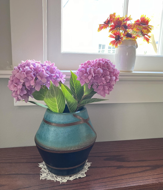 In the left foreground is a black and turquoise vase made from stoneware--it is holding two bright pink hydrangeas. In the upper right background is a small white vase sitting on a window sill--it is holding a bunch of red and yellow Coreopsis wildflowers. 