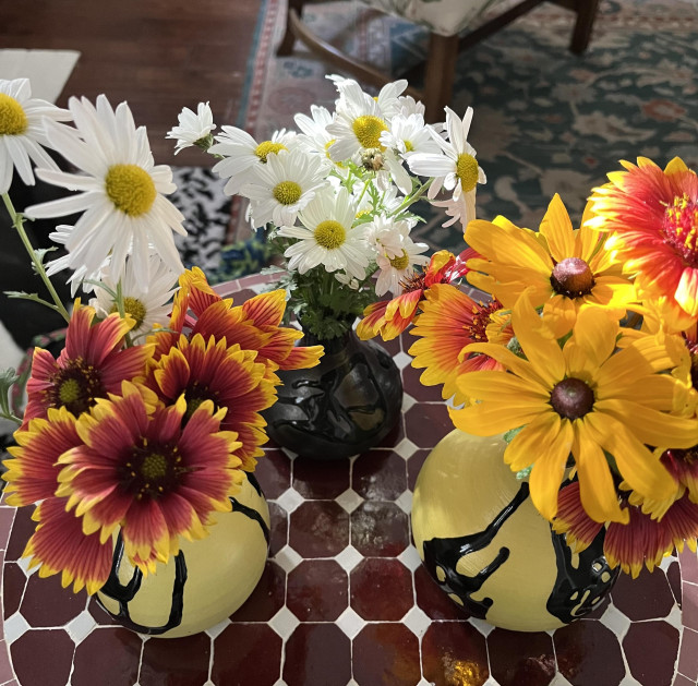 Three vases holding a mixture of red and yellow Coreopsis wildflowers and an old variety of white Chrysanthemums. Two vases are yellow with black drips, and the third vase is black with black drips.