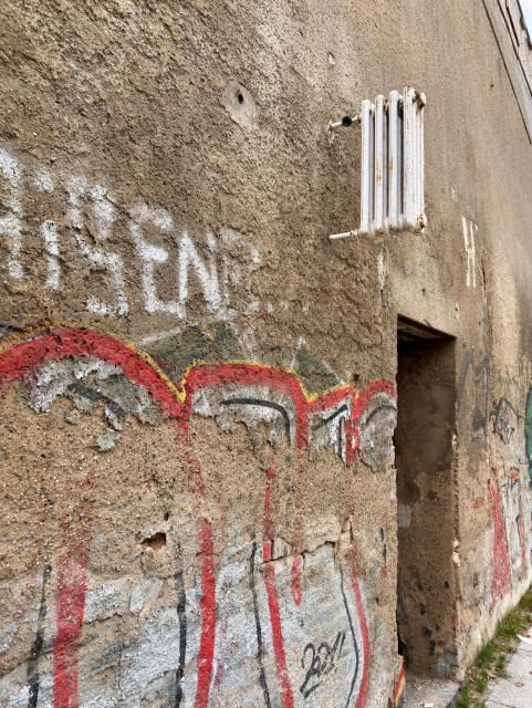 Eine verwitterte Wand mit strukturierter, abblätternder Farbe und Graffiti. Ein weißer Heizkörper ist oben montiert, und auf der rechten Seite befindet sich eine Tür. Das Graffiti zeigt rote und schwarze Designs, mit einigen weißen Schriftzügen, die teilweise sichtbar sind.