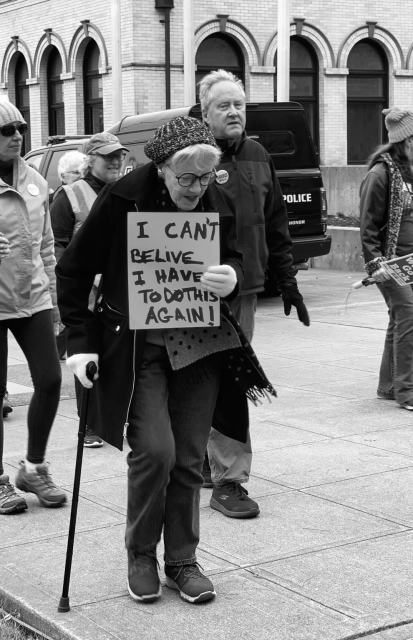 Black and white photo of older lady with cane marching with handwritten sign "I can't believe I have to do this again!"