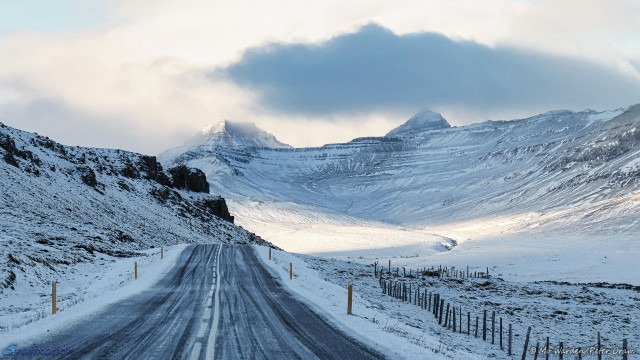 A snow scene, viewed from inside a car on an icy road that is about to crest a small hill. There's a fence line following the side of the road for a short distance and on the left is a rocky hillside. Both sides of the road are lined by tall snow-poles. The sky is grey and full of heavy cloud, but there is sunlight from the left of the shot. Ahead are two prominent mountain peaks with a long glacial valley curving down, with a water-cut gorge between them. It almost looks as if the road continues over the hill and up the side of the mountain.