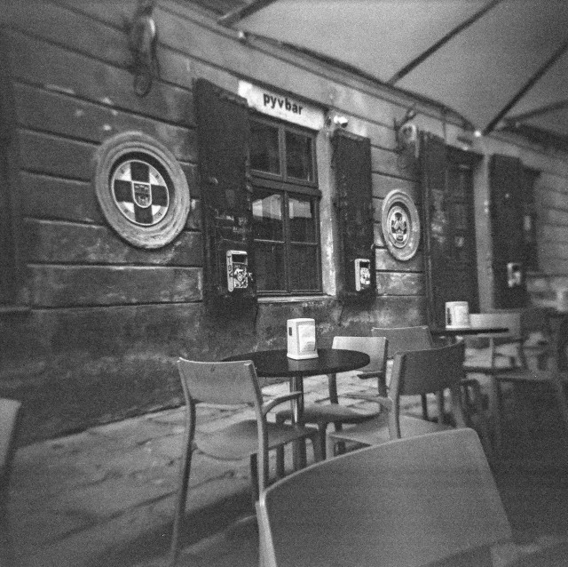 This image shows an outdoor seating area in front of an old building, likely a bar or pub. The scene is in black and white, giving it a nostalgic or vintage feel. The building has weathered, textured walls with wooden shutters on the windows and some circular emblems or signs mounted on the wall. Above the window, there's a small sign that reads "pyvbar." The tables and chairs are simple and modern, with square napkin holders placed on the tables. The space appears quiet and empty, possibly in the early morning or late evening when the area isn't crowded. The overall mood is calm