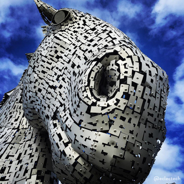 A square photo of one of The Kelpies. It is a large steel sculpture of a horse head. This photo is taken standing just underneath the one that has its head down, so its snout is very close to the camera and it appears to be smiling down at the photographer. Behind it is a bright blue sky with wispy white clouds.