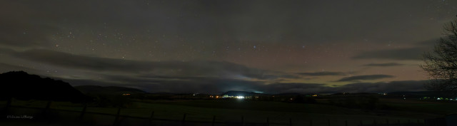 A wide night sky panorama soloing a low dark horizon with the lights of houses scattered along it. A hill is highlighted and pale white rays of vehicle lights can be seen coming from behind that hill, as the eye moves right more colours appear as a soft Aurora displays greens and shades pinks \ reds. A tree is silhouetted on the far right