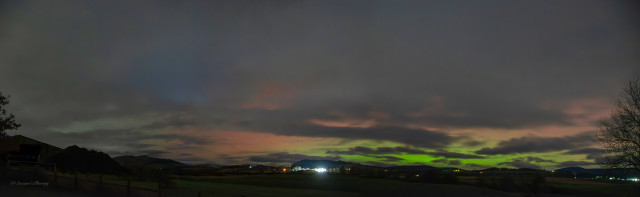 A wide night sky panorama soloing a low dark horizon with the lights of houses scattered along it. A hill is highlighted and Aurora colours can be seen above that hill, as the eye moves right more colours appear as a bright Aurora displays greens and shades pinks \ reds. A tree is silhouetted on the far right