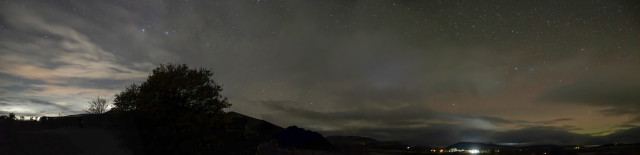 A wide night sky panorama soloing a low dark horizon of distant trees to the far left, a fence and shrub as the eye moves right to a  silhouetted tree. The sky behind all that is streaked with light of various shades, whites, pale pinks and greys caused by the lights of the town. To the right of centre the Aurora starts to show through streaks of dark clouds, some lit up by vehicle lights, to the far right are the orange, pinks, greens of the Aurora