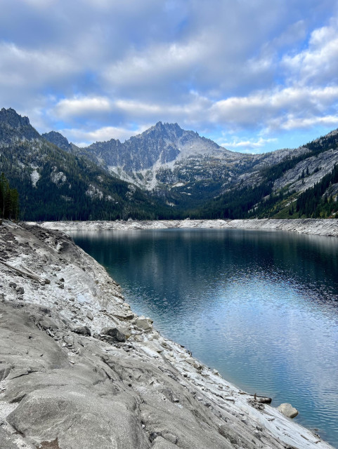 A view of Prusik Peak rising above upper Snow Lakes in The Enchantments. The lake is surrounded by several feet high gray rock banks. The peak is jagged dark gray rock. The sky is full of gray and white clouds with blue sky showing through. Photo taken September 22, 2023, near Leavenworth, Washington, U.S.