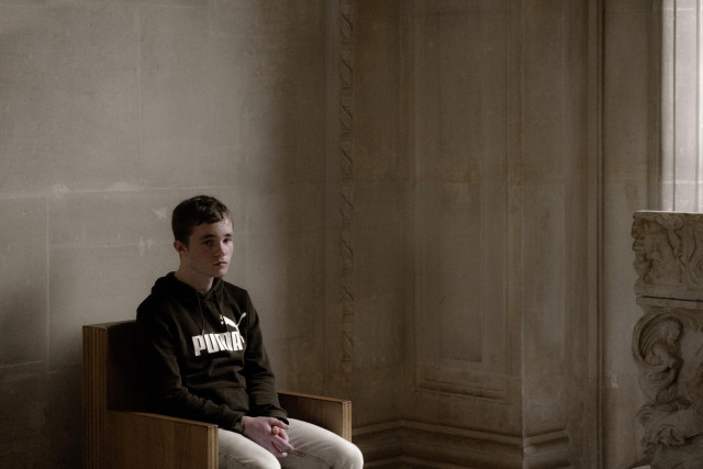 A young person sits alone in a dimly lit, austere room, gazing thoughtfully toward the side. They are dressed casually in a black Puma hoodie and light-colored pants, contrasting with the classical stone walls and an ornate carved stone pedestal nearby. The setting has a contemplative, almost melancholic feel, with the muted lighting enhancing the somber atmosphere.