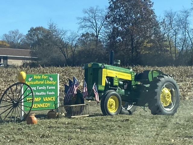 A vintage John Deere tractor is positioned in a field, accompanied by a sign that promotes agricultural liberty and mentions candidates. The sign is decorated with small American flags and pumpkins, set against a backdrop of trees and a clear blue sky.