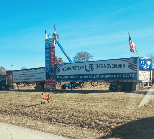 A large billboard on trucks promotes voting, emphasizing the message "PLEASE VOTE FOR LIFE THIS NOVEMBER." There are banners with religious themes and political statements, including references to good versus evil and the importance of prayer.