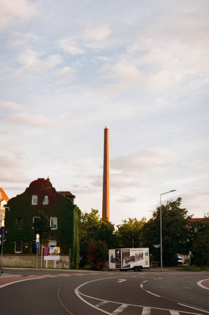 The analog photo shows an ivy-covered building by a curved road, with a tall brick smokestack rising prominently in the background against a lightly clouded sky. A trailer with advertisements is parked nearby.