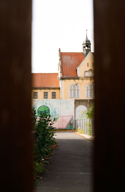 Seen through a narrow gap, an old building with a red-tiled roof and small tower stands behind a graffiti-covered wall (one prominent graffiti reads “laut!”, which means “loud”).