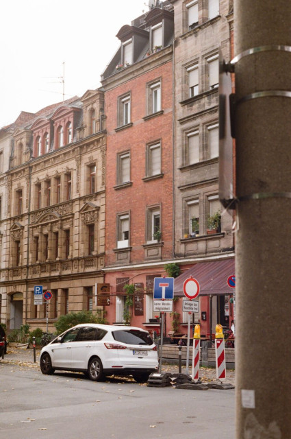 The analog photo shows an empty street with buildings with old facades/brick structures and in the middle a parked white car in front of mobile street signs.