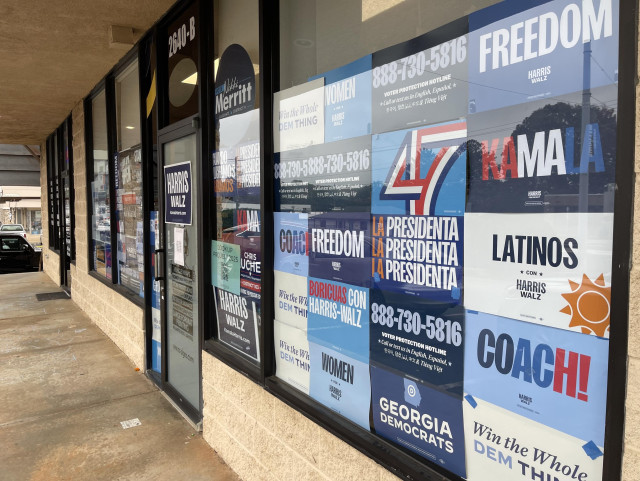 The image features a storefront with large windows displaying various political campaign signs. The signs promote "Harris Walz," "Kamala," and messages related to freedom, democracy, and voter protection. There are references to women, Latinos, and Freedom. 