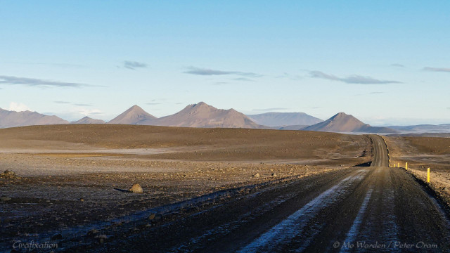 A photo of a landscape under an almost completely clear cyan sky. On the horizon is a range of bare mountains, steep-sided brown rock. The foreground is an undulating plain of brown dirt, covered in scattered rocks sized from gravel to boulders. From bottom centre to middle right of the shot, a dirt road lined on one side by snow-poles is snaking ahead and disappearing over a rise. Beyond it, flat ground can be glimpsed.