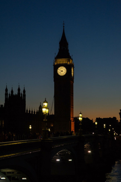 Late evening photo of a tall square tower with a pointed roof just under which are internally illuminated clock faces on each side. To the left is a silhouette of another large square tower with smaller ornate towers on each corner. Before both of these is a bridge crossing a river which is sky-lit in the bottom-right corner by the orange-blue darkening sky. The arches under the bridge are lit from the inside and across its top are two sets of old-fashioned streetlights and many pedestrians, just about visible in the dark.