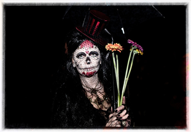 A portrait of a young woman in Day of the Dead makeup and costume. White and red skull face, black veil and dress, black fingernails. She holds 3 long-stemmed flowers.