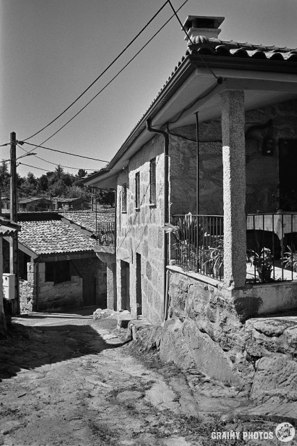 A black-and-white film photo of stone houses by a narrow road. The first house on the right has a large covered terrace nearest the camera.