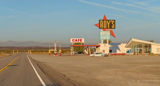 Color landscape photo of a deserted two-lane highway on the left. On the right is a large sign that says, "Cafe," over a service station awning. Further to the right, and in front of the service station, is a much larger sign that reads, "Roy's vacancy motel cafe." That sign has a red triangular wedge that points down at the motel office. "Roy's" has a black background and "motel cafe" is on a blue background. The motel office has a wedge shaped roof that contrasts with the red triangular sign above it. 